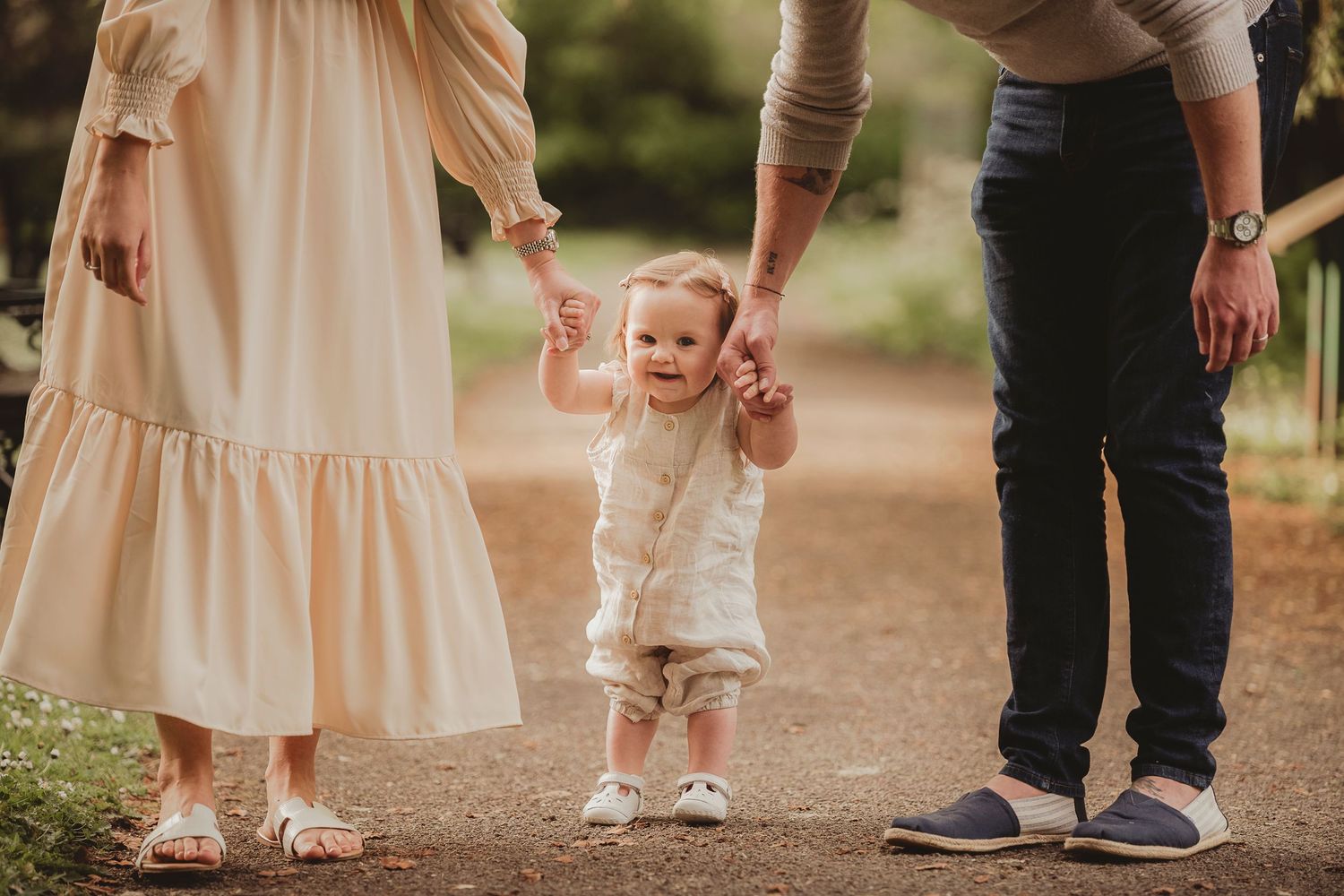 Small child taking first steps while holding parents' hands on outdoor path.