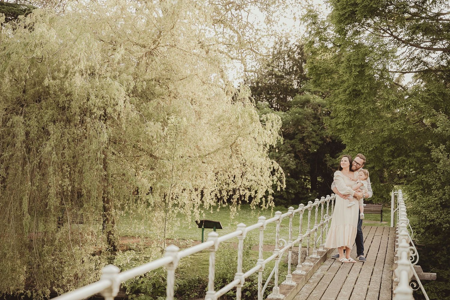 Romantic scene on wooden bridge with willow trees and white fence in background.