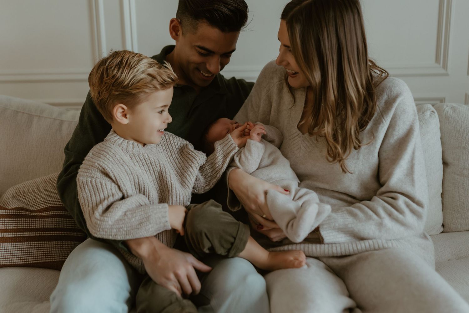 A family sits together on a white bed sharing a tender moment in a cozy bohemian bedroom with neutral decor.