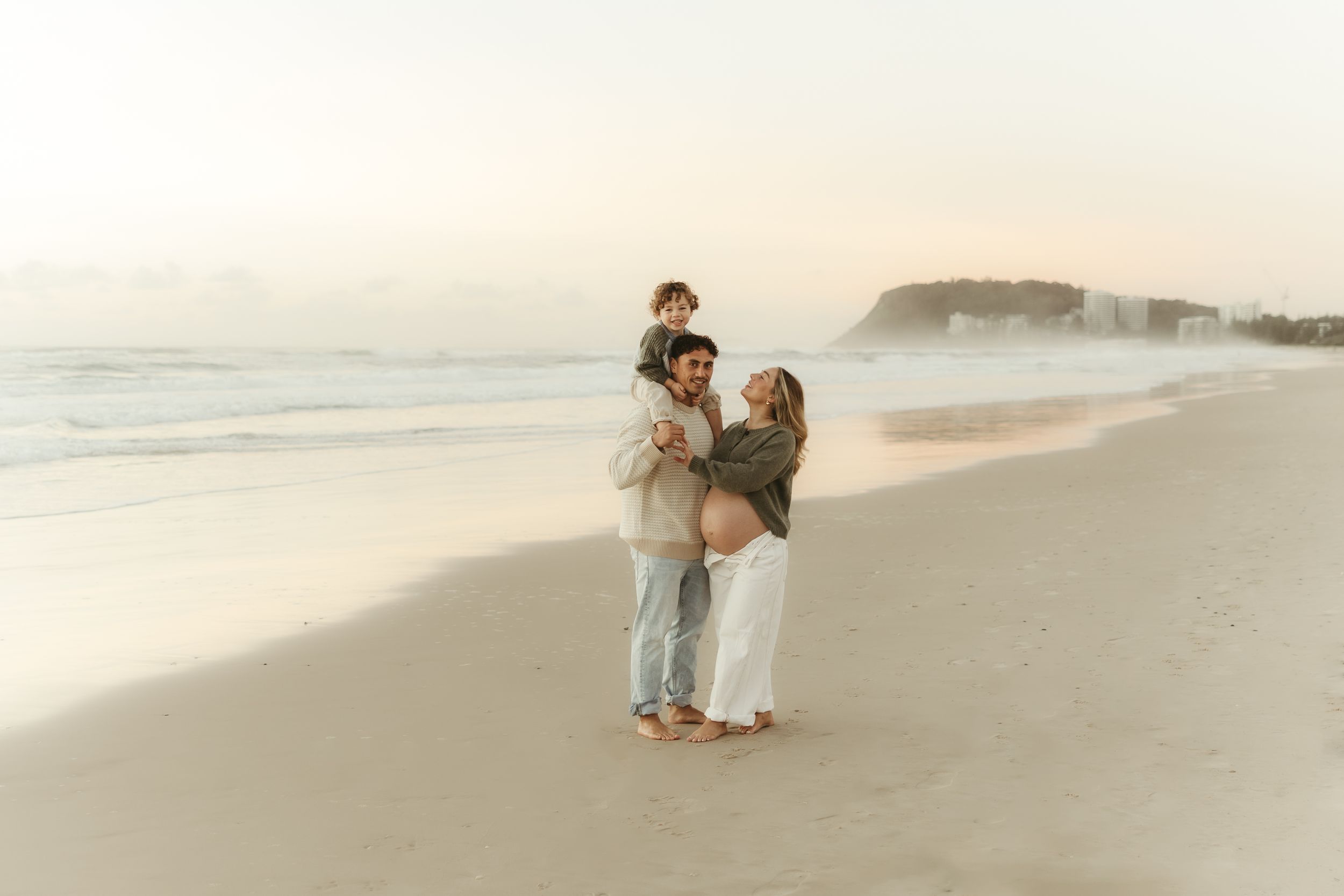 Pregnant couple with toddler on shoulders sharing a tender beach moment at sunset on a sandy shore.