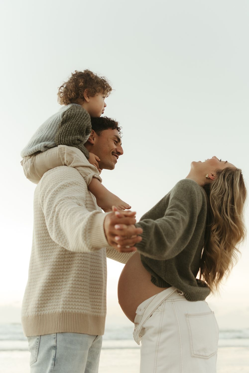 Happy family at beach, pregnant woman and man with toddler on shoulders, enjoying sunny day outdoors.