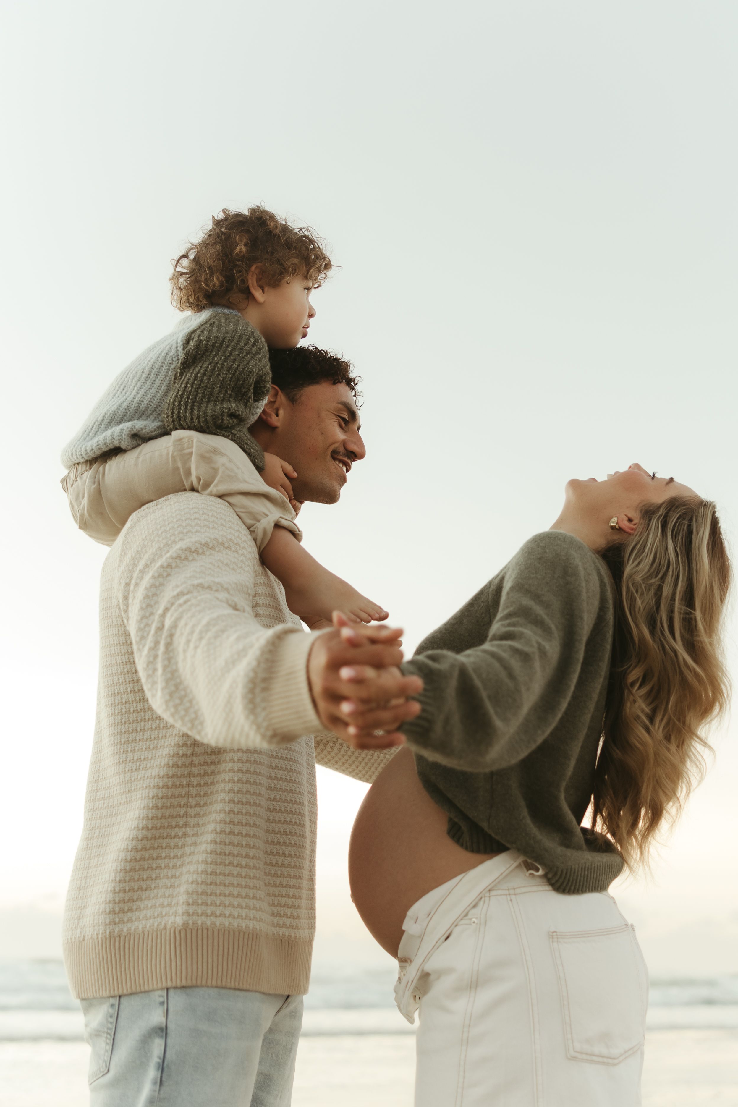 Happy family at beach, pregnant woman and man with toddler on shoulders, enjoying sunny day outdoors.