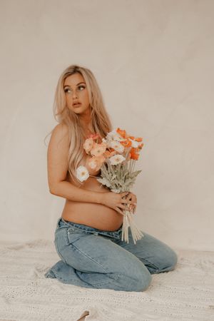 A person in light blue jeans sits on a beige background holding flowers in a sensual maternity boudoir photoshoot.