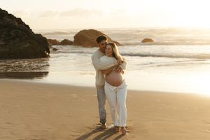 Couple embracing on sandy beach at golden sunset, pregnant woman in white outfit, ocean waves and rocks in background.