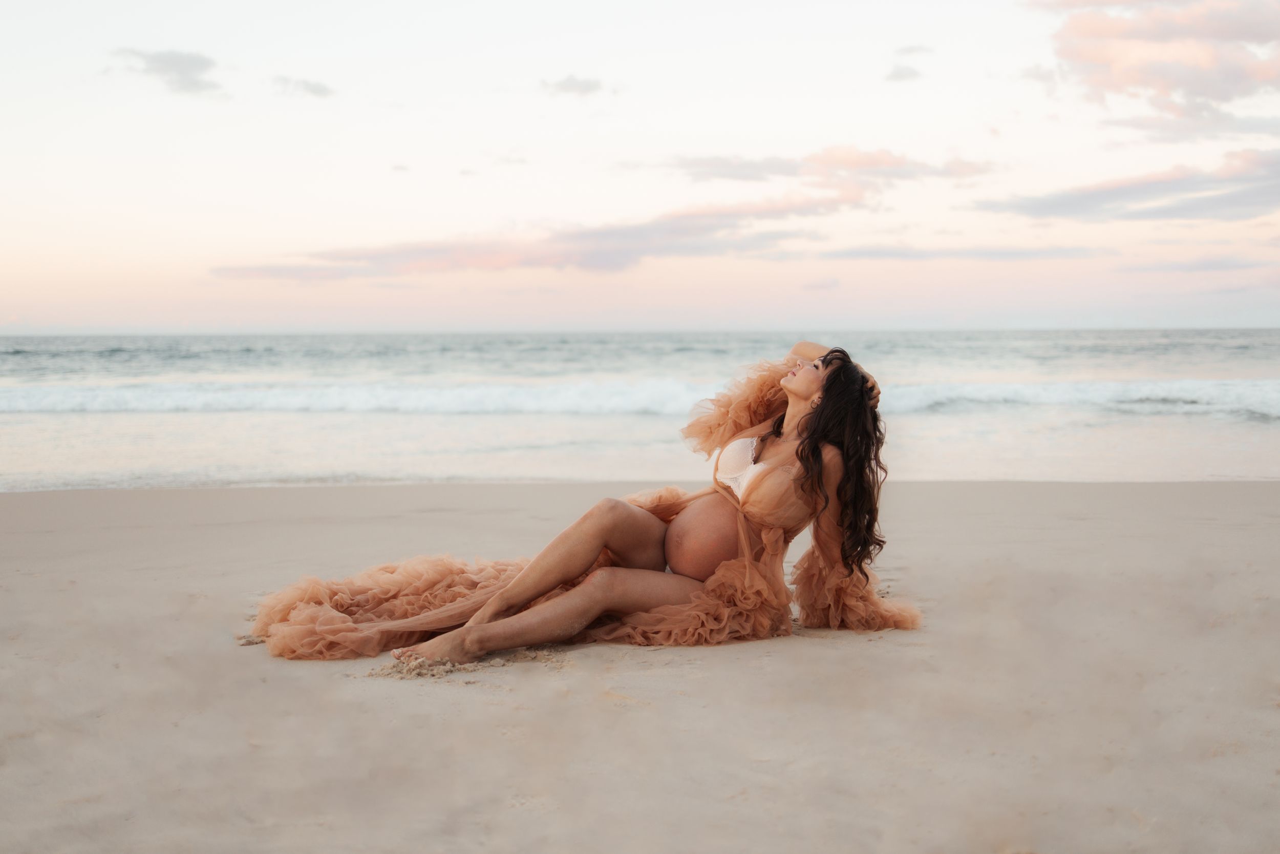 A serene beach portrait features flowing peach fabric against the ocean at sunset.