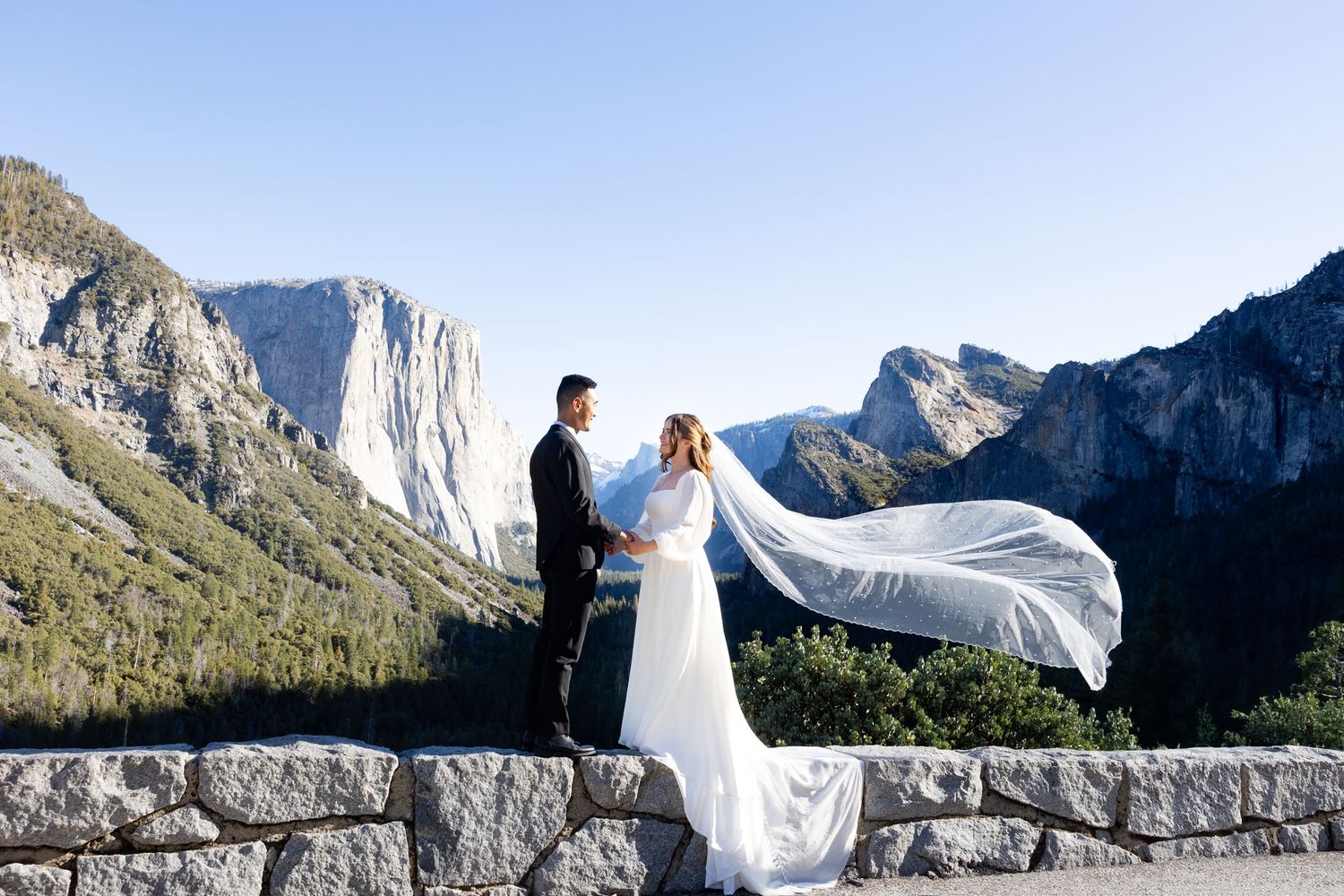 A couple in wedding attire stand at Tunnel View overlook with dramatic El Capitan and Yosemite Valley cliffs in background.