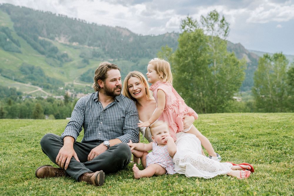 Happy family of four sitting on green grass with scenic mountain views in background on a cloudy day.