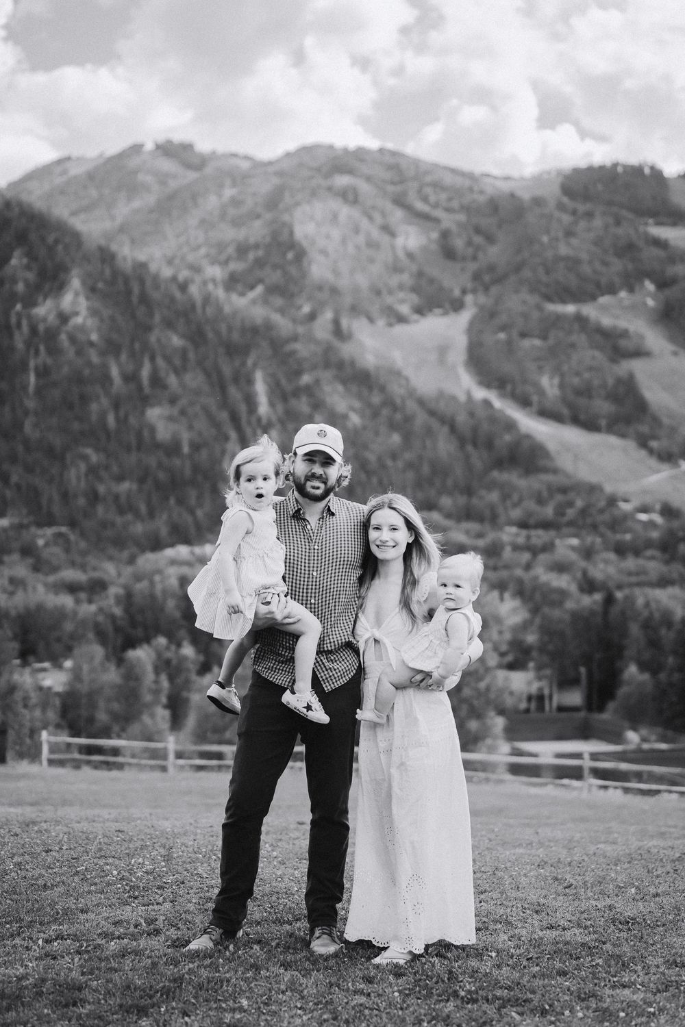 Black and white family photo with parents holding two young children, posing in a mountain meadow with forested peaks behind.