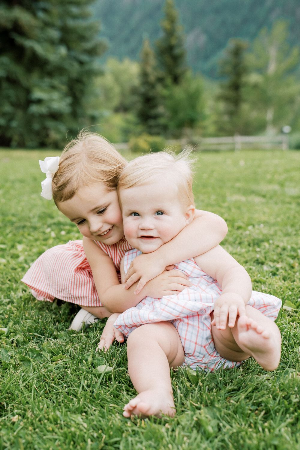 Two young siblings hugging on green grass with mountain trees backdrop, older girl in pink dress embracing smiling baby.