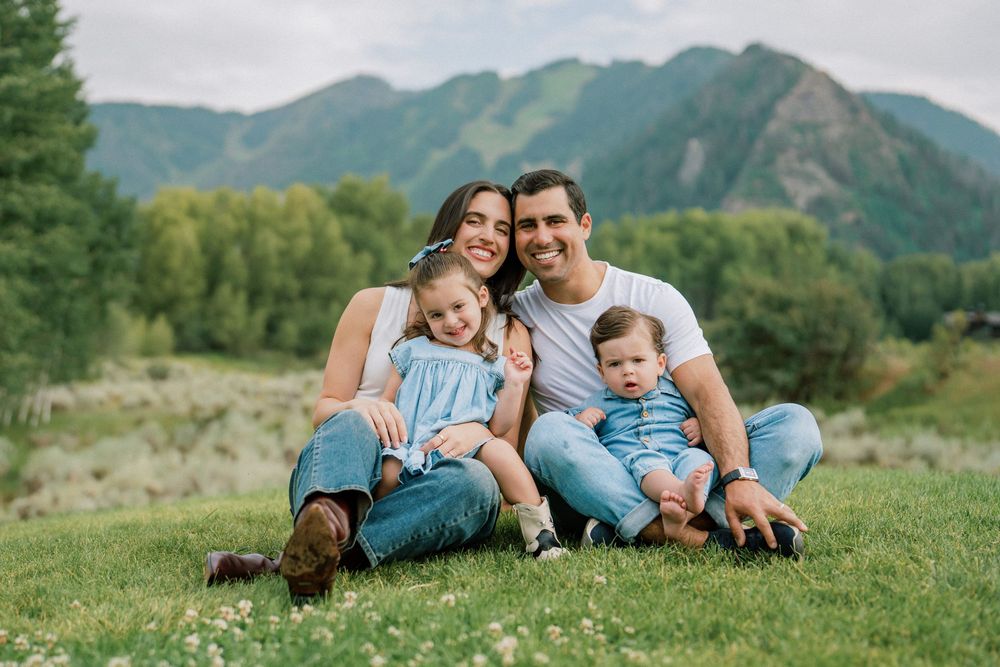 A family sits together on a grassy hillside with mountains and trees in the background on a sunny day.