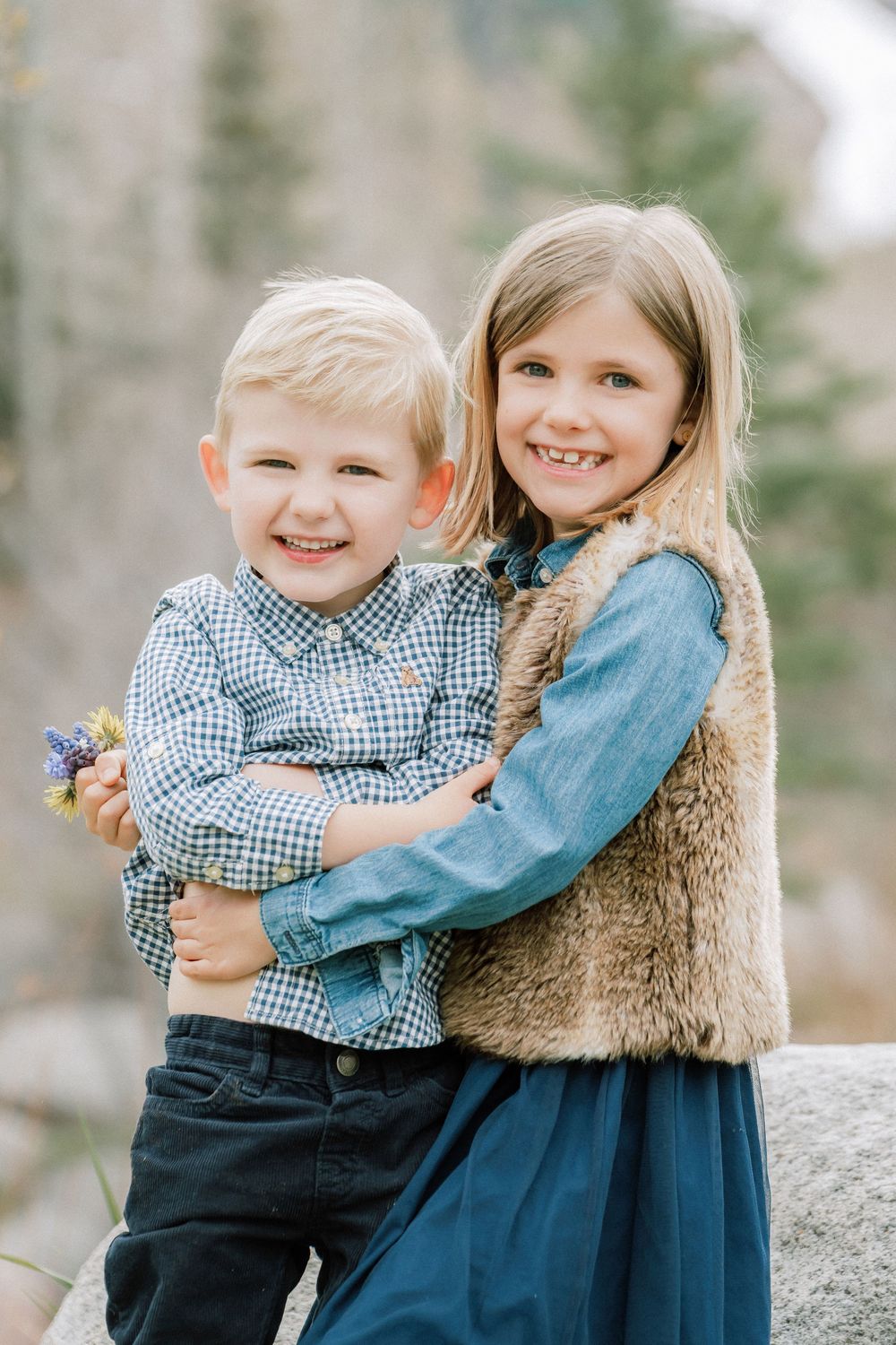 Two smiling children pose outdoors, a boy in plaid shirt holding flowers hugged by a girl in denim and fur vest.