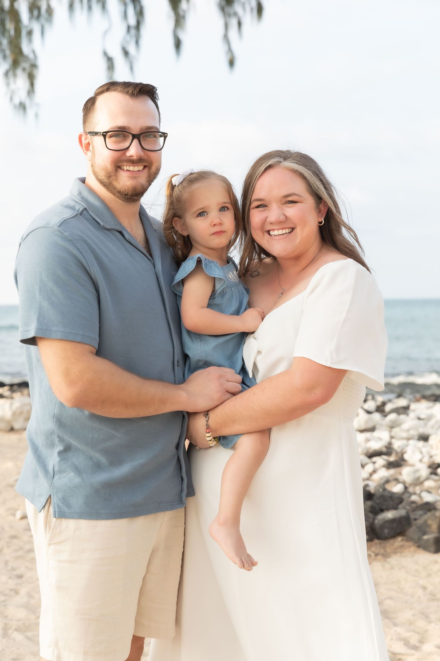 Happy family in coordinated blue and white outfits pose together on a tropical beach with palm trees and ocean backdrop.