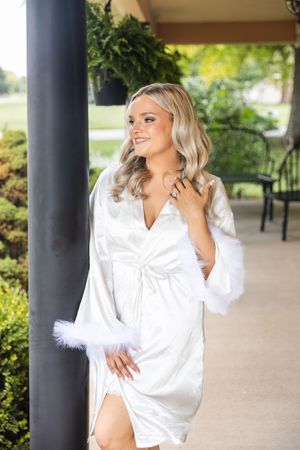 Bride posing in white silk robe with feather trim poses on outdoor porch in New Virginia, Iowa. Wedding photography ideas for brides.