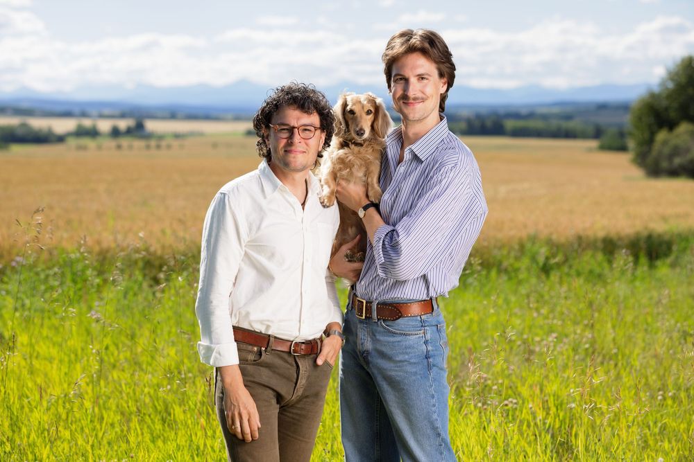 Two men smiling outdoors in green field holding a golden cocker spaniel dog.