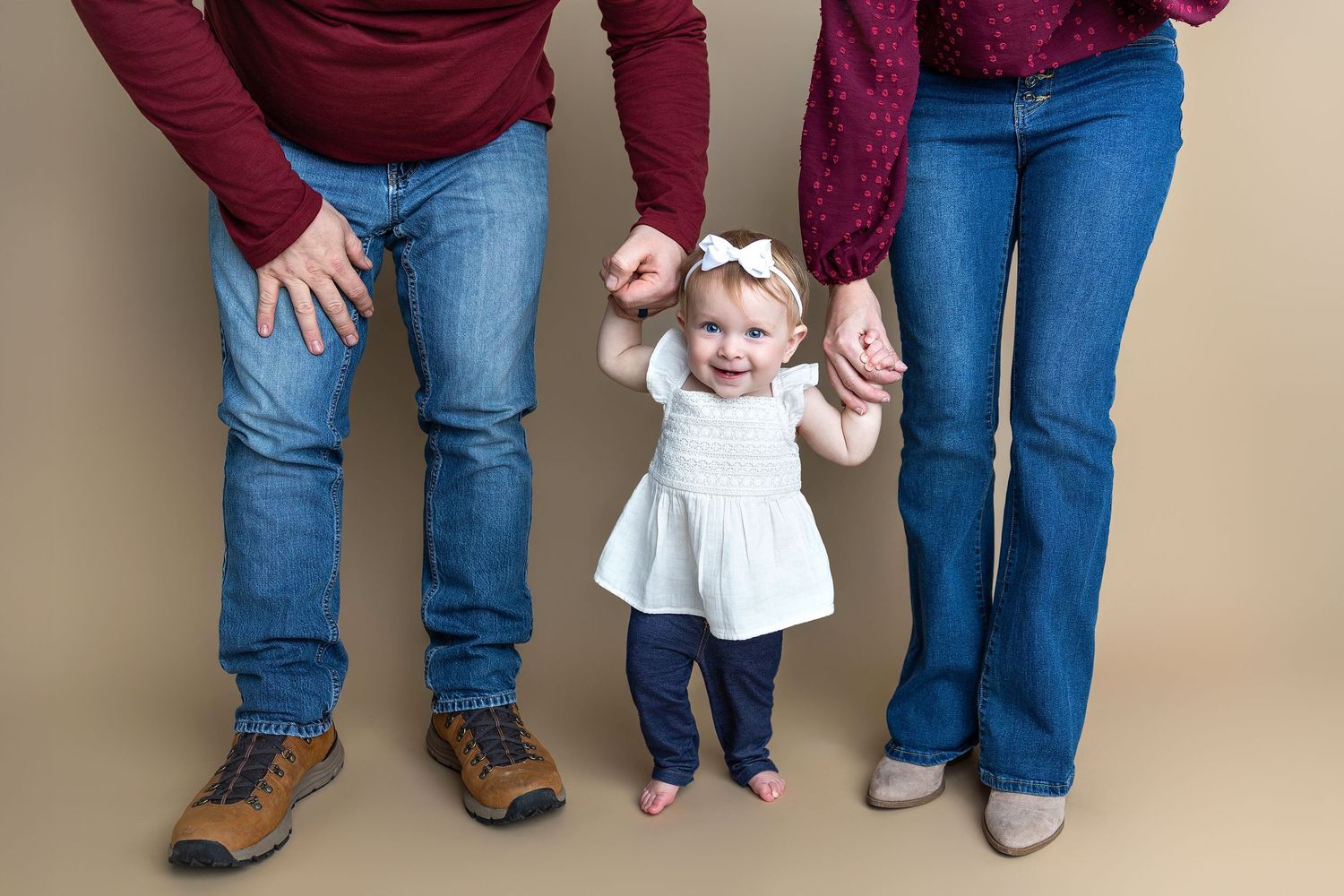 Parents holding hands with toddler learning to walk on grass.