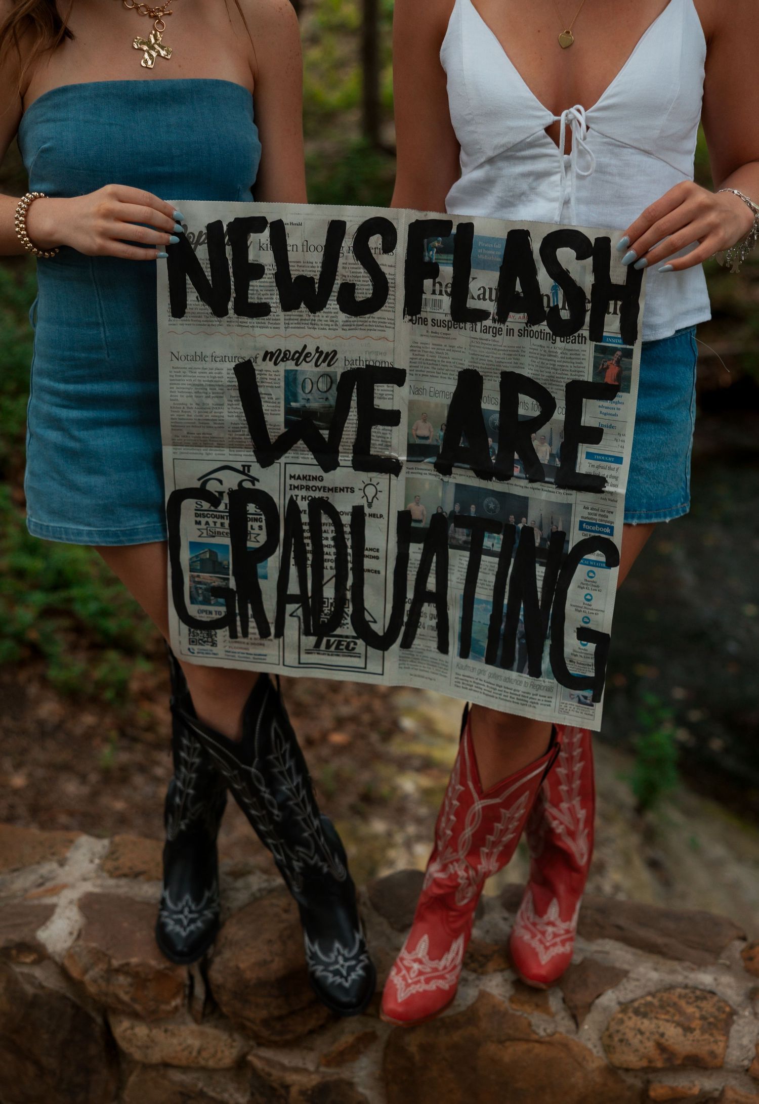 Two graduating women in denim skirts and cowboy boots holding a newspaper graduation sign.
