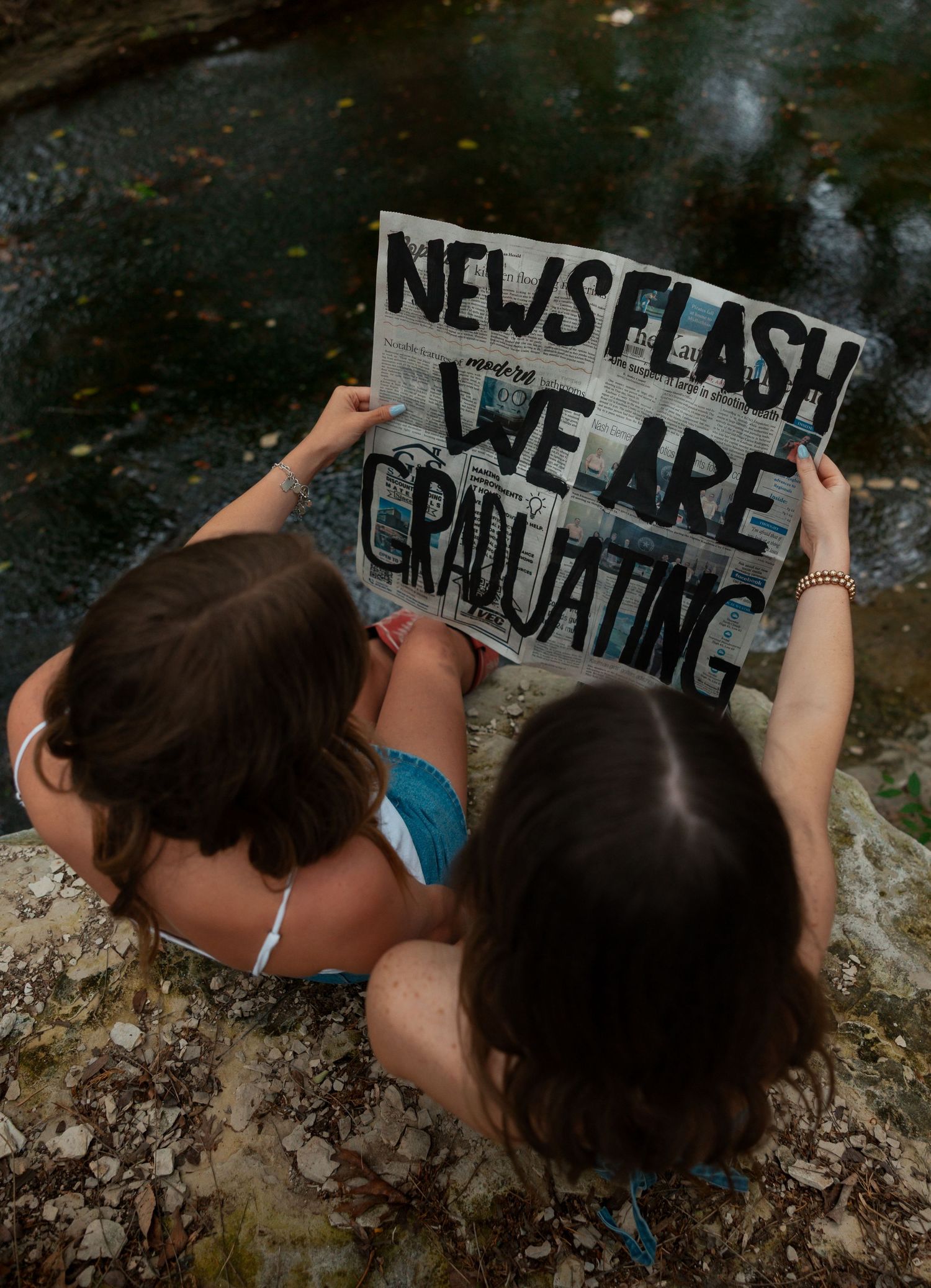 Two young women sitting in a creek holding a News Flash We Are Graduating newspaper sign.
