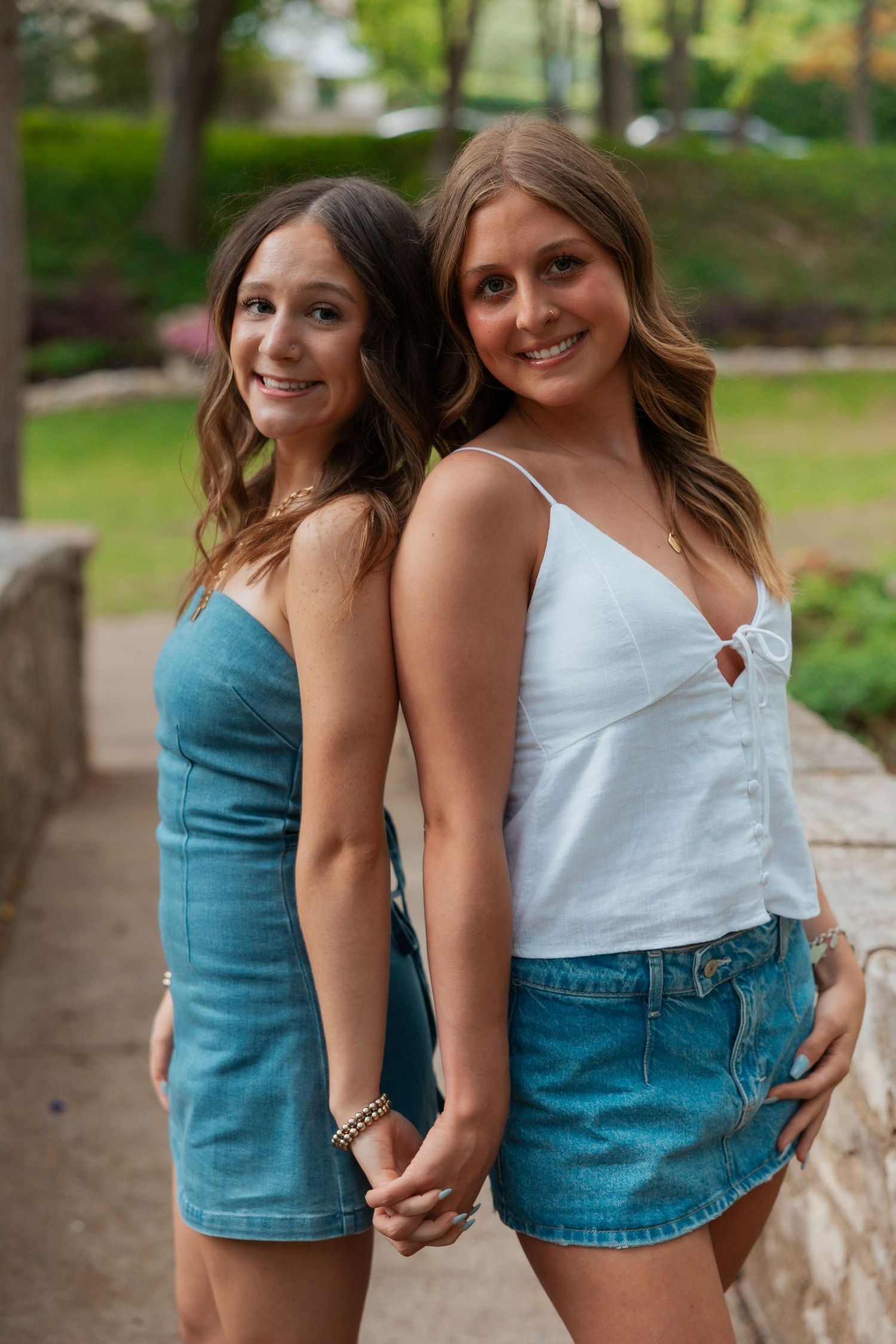 Two smiling young women in denim outfits posing back to back on a stone walkway outdoors.