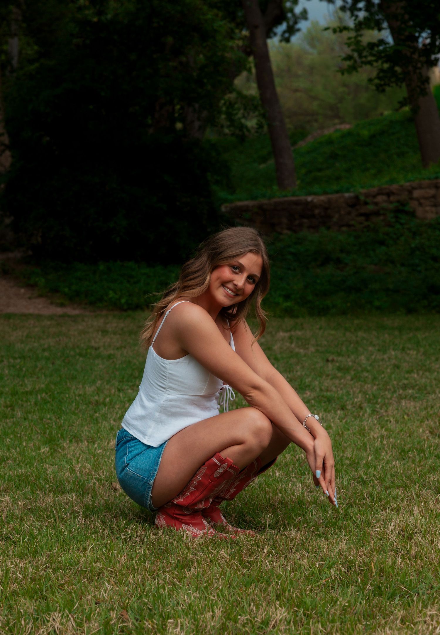 Young woman in white tank top, denim shorts, and red cowboy boots crouching on green grass outdoors.