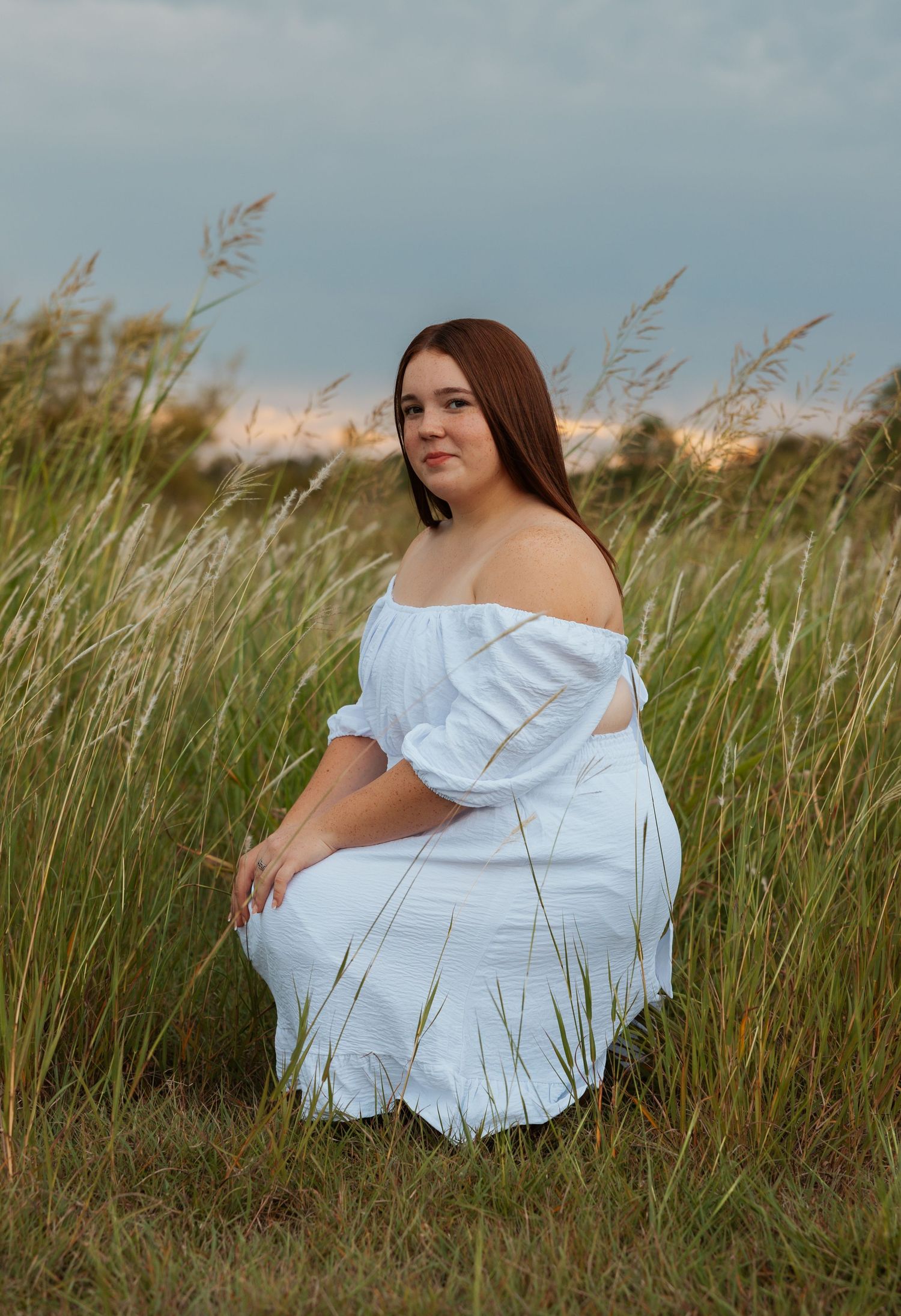Young woman in white off-shoulder dress sitting gracefully among tall grass at golden hour.