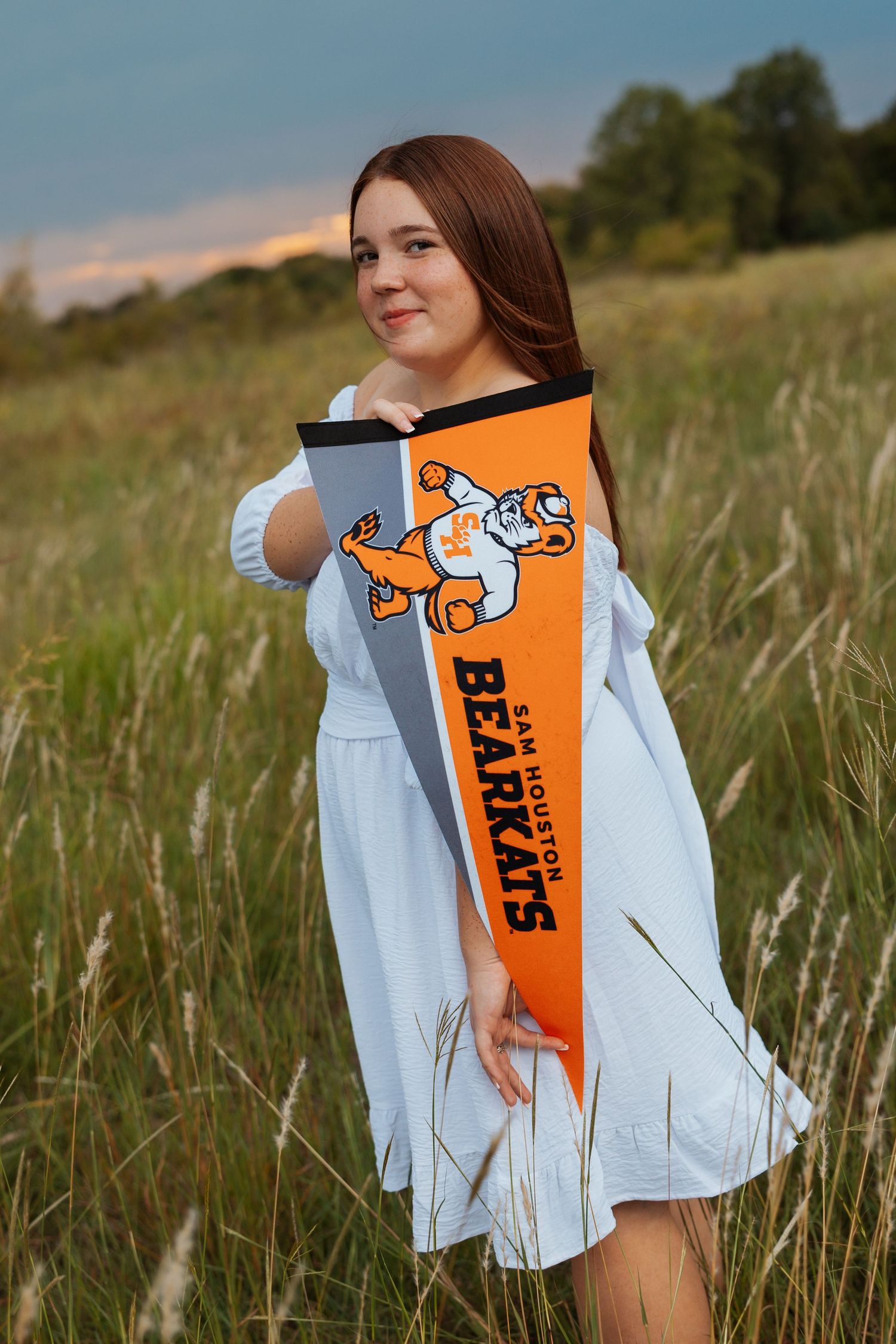 Young woman holding Sam Houston Bearkats pennant in a field at sunset wearing a white dress.