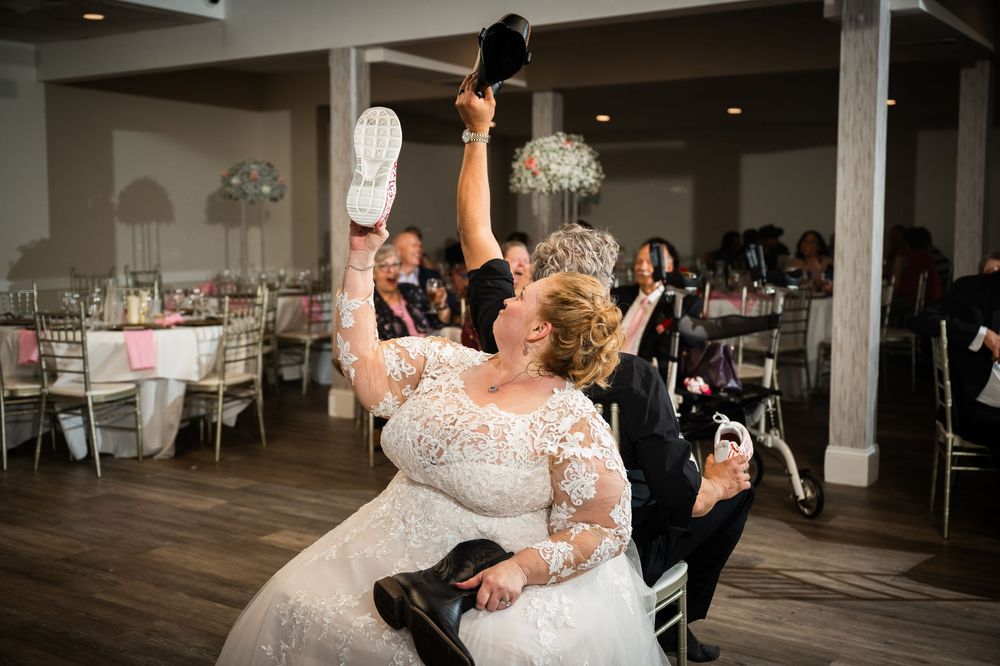 Bride in lace wedding gown holds up shoes during fun wedding reception shoe game at elegant venue.