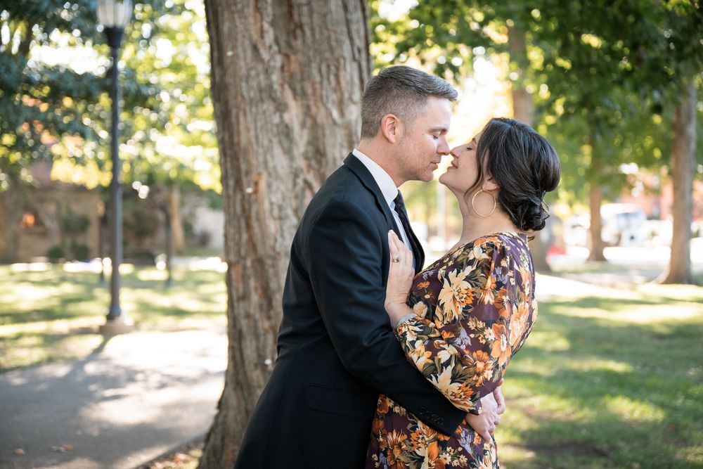 A couple shares a romantic kiss in a sunlit park setting with tall trees in the background.