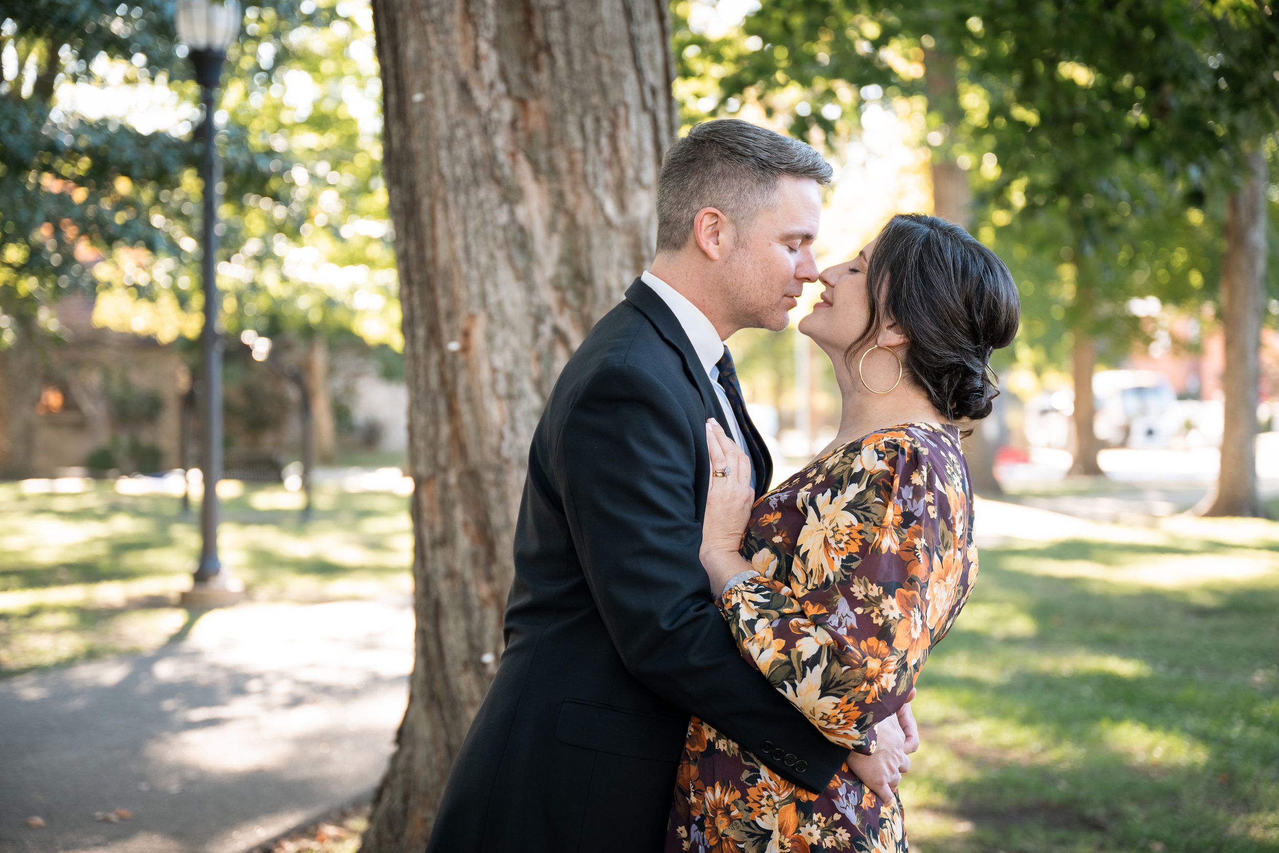 A couple shares a romantic kiss in a sunlit park setting with tall trees in the background.