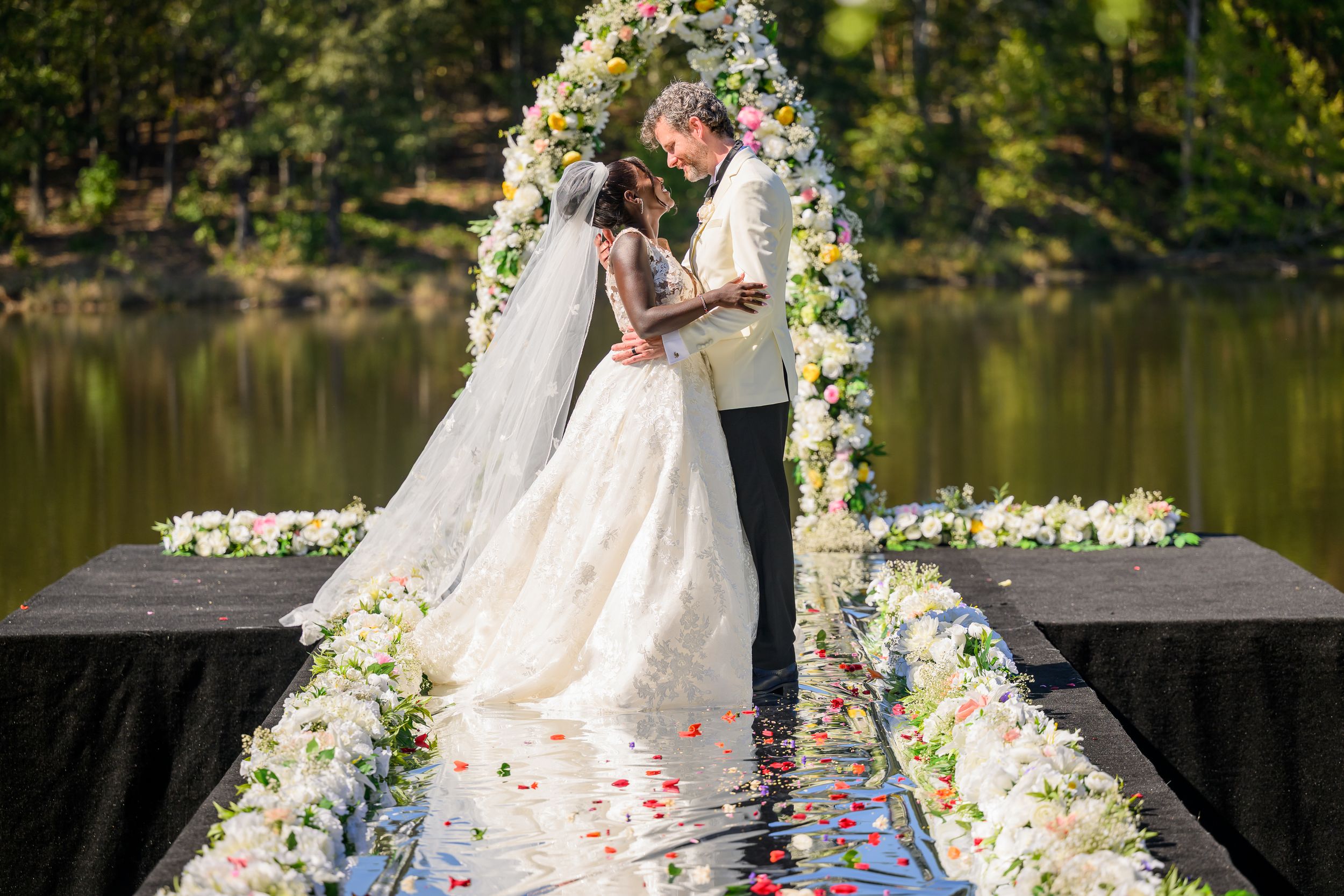 Wedding ceremony setup with floral arch and rose petals along a lakeside walkway.