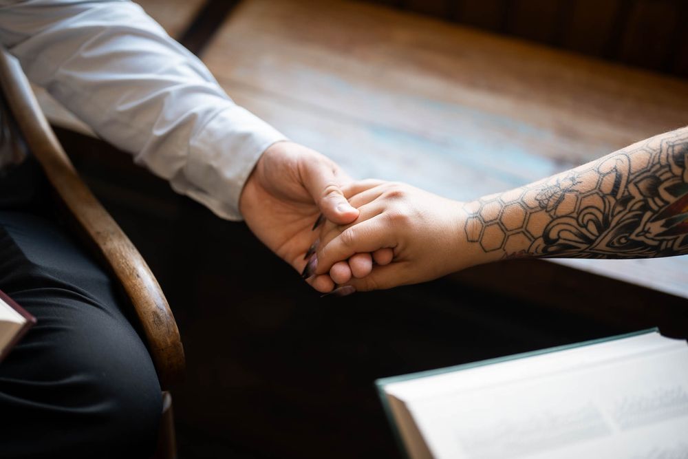Two people holding hands, one with a floral sleeve tattoo, sitting together at a wooden table.
