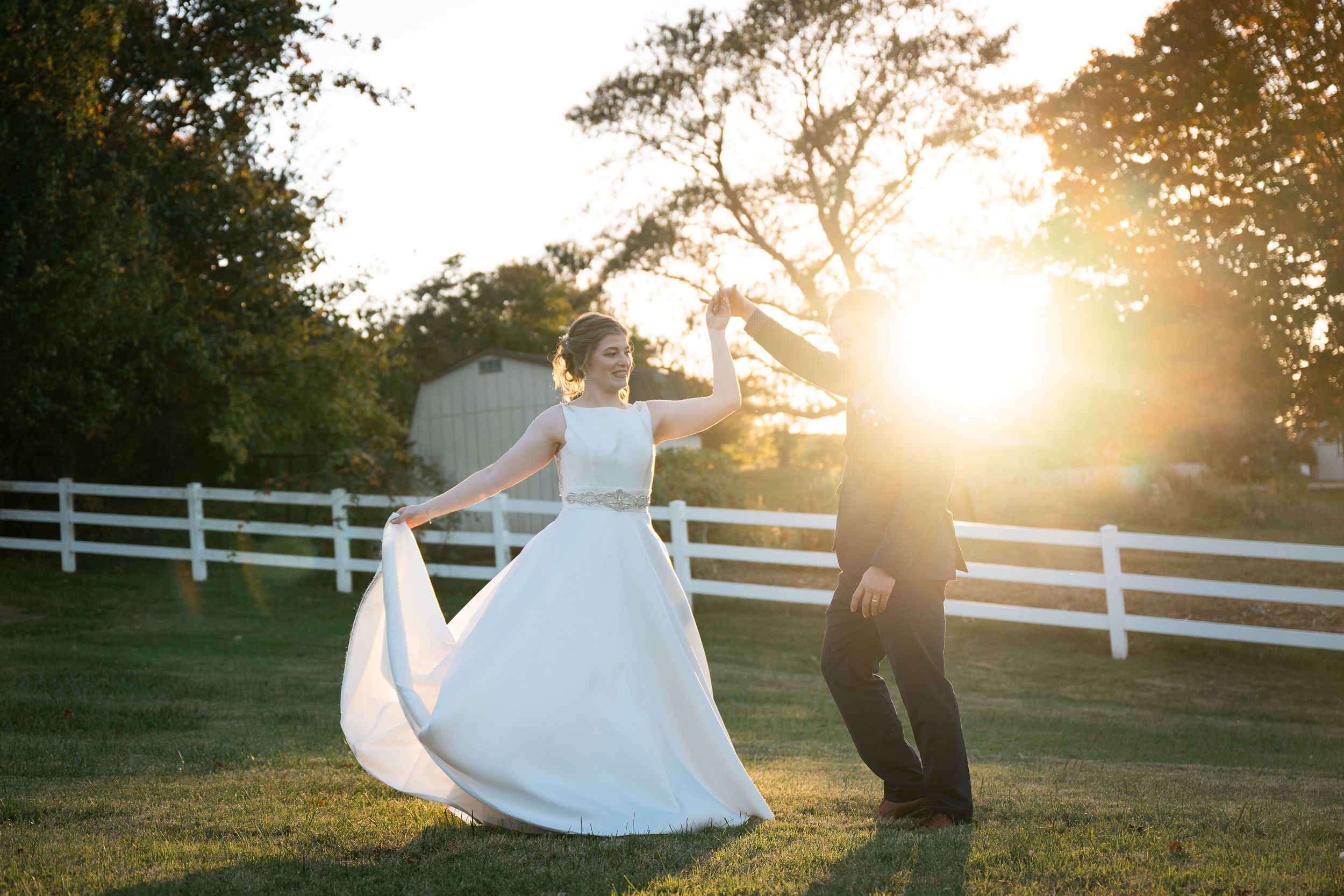 A couple dances playfully at sunset with white fencing and trees in the background.