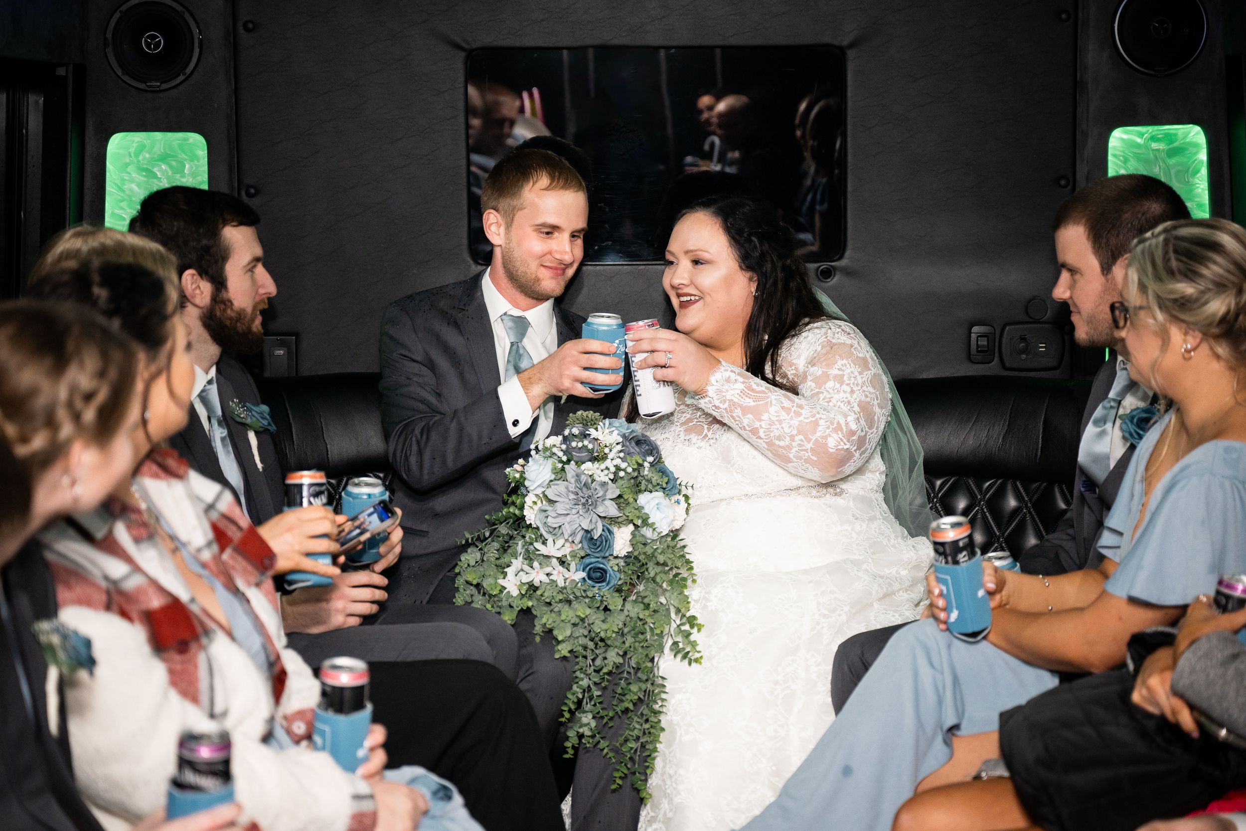 Wedding party celebrates together in a dimly lit party bus with blue drinks and greenery decorations.