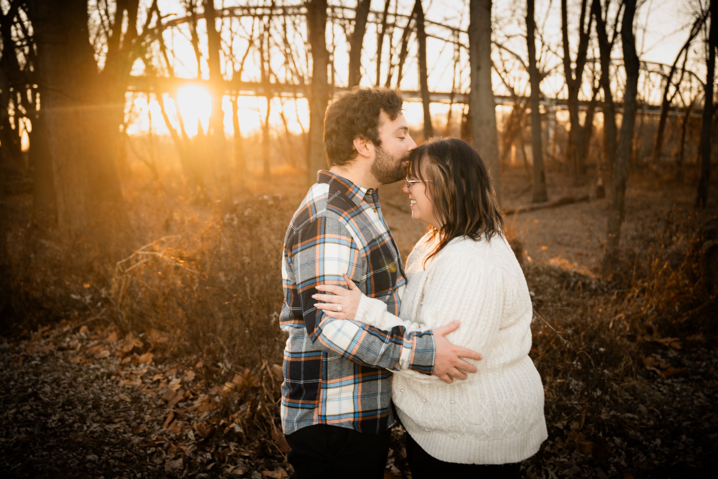 Couple sharing a romantic moment at sunset in a wooded area, golden light glowing behind bare winter trees.