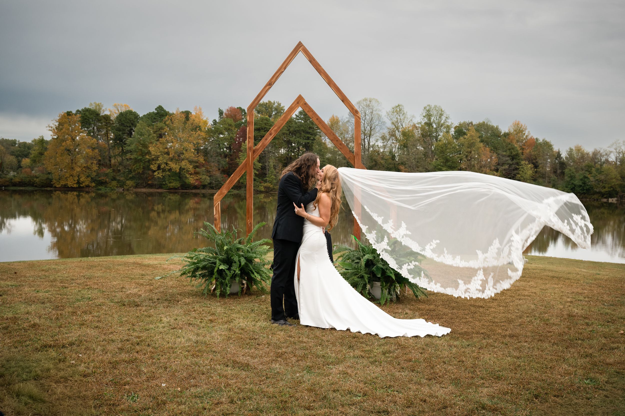 A couple in formal wedding attire poses together in a grassy field with a flowing veil caught in the breeze.