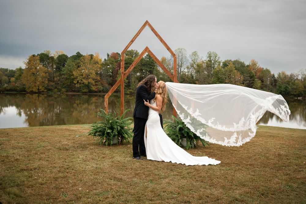 A couple in formal wedding attire poses together in a grassy field with a flowing veil caught in the breeze.