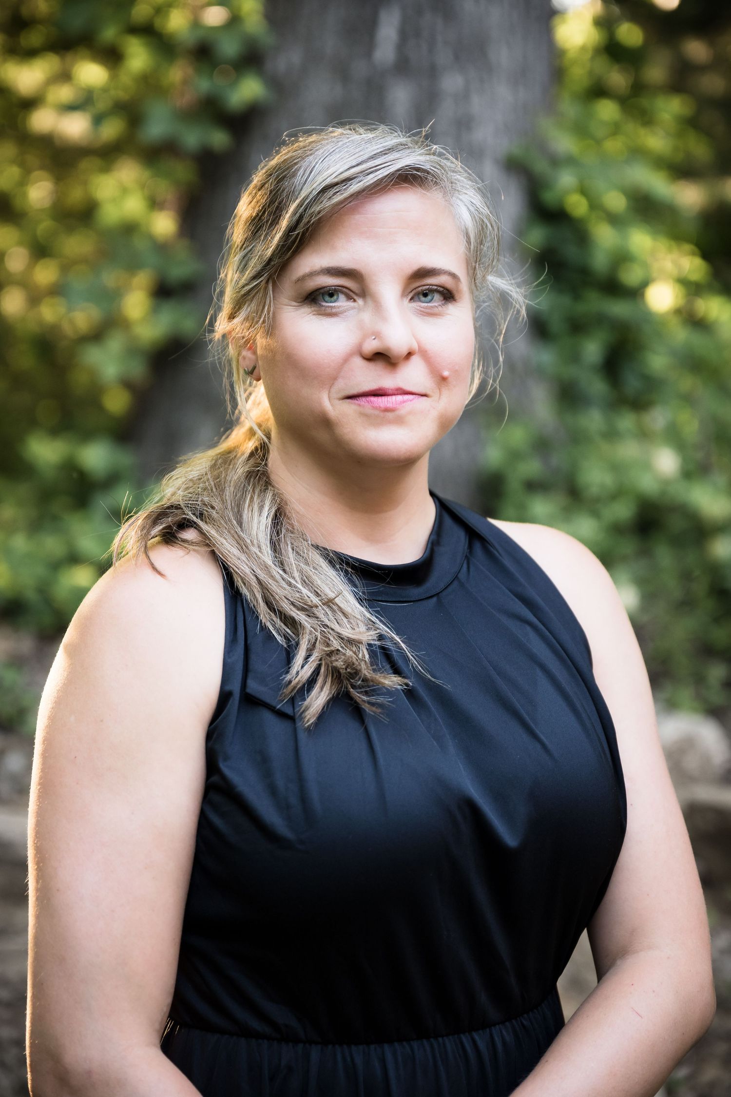 A person wearing a sleeveless black dress poses outdoors with natural greenery in the background.