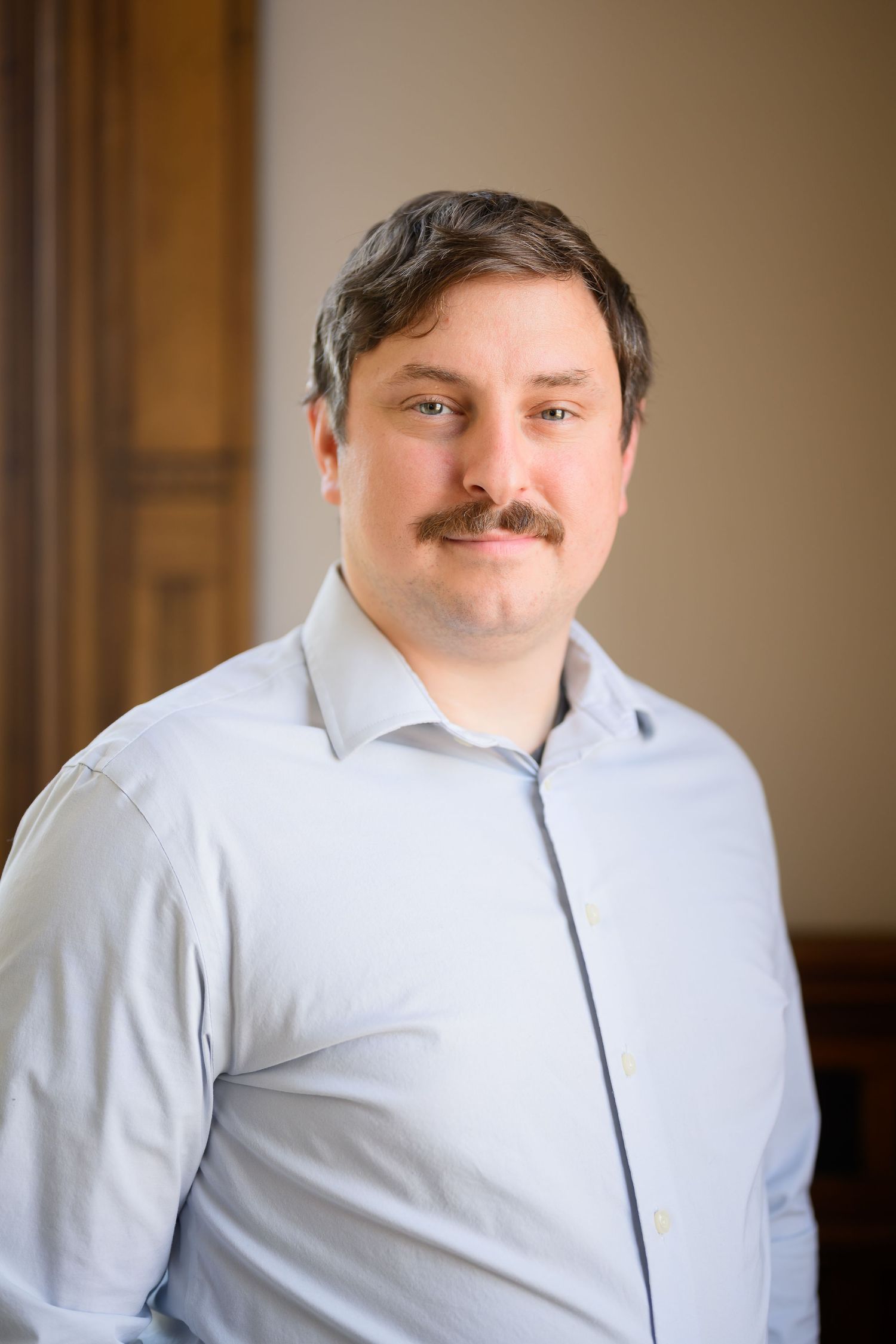 A person wearing a white dress shirt smiles warmly at the camera in a professional indoor setting.