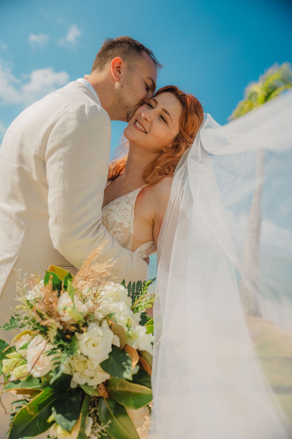 Romantic beach wedding portrait with white flowers and flowing veil against bright blue sky.