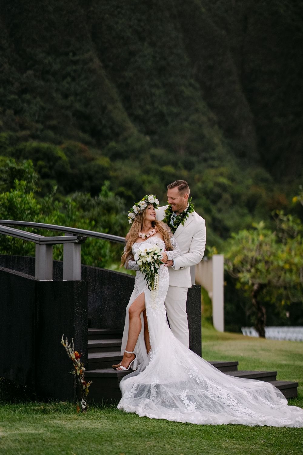 Bride and groom embrace on steps with lush green Hawaiian mountains backdrop, bride in lace gown with floral crown.