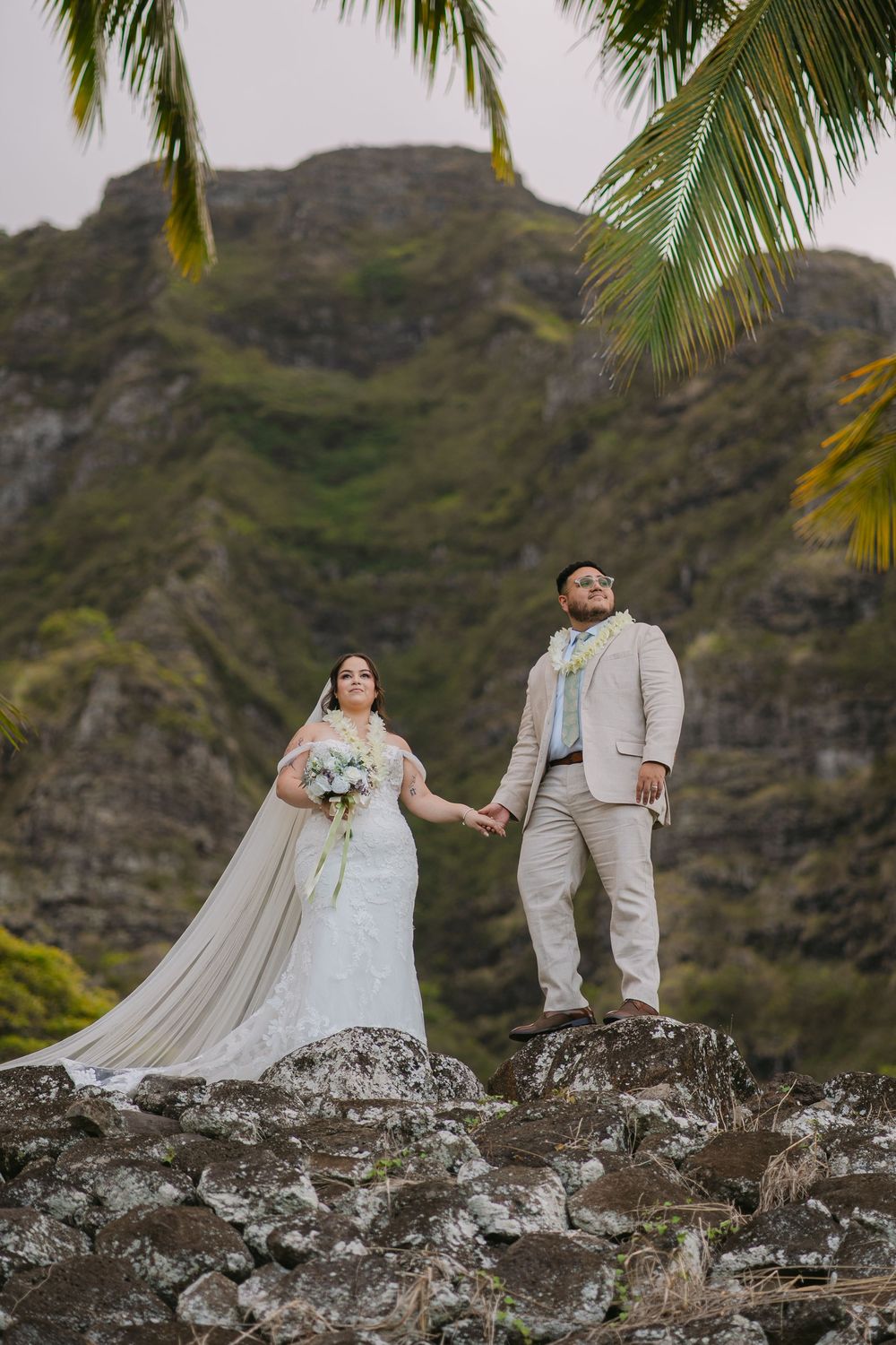 A couple in wedding attire stands on rocky cliffs with palm trees and mountains in the tropical Hawaiian backdrop.