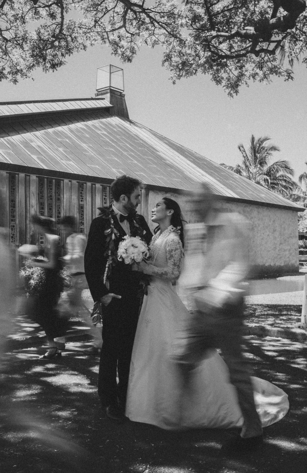 Vintage black and white wedding photo outside a rustic building with dappled sunlight through trees.