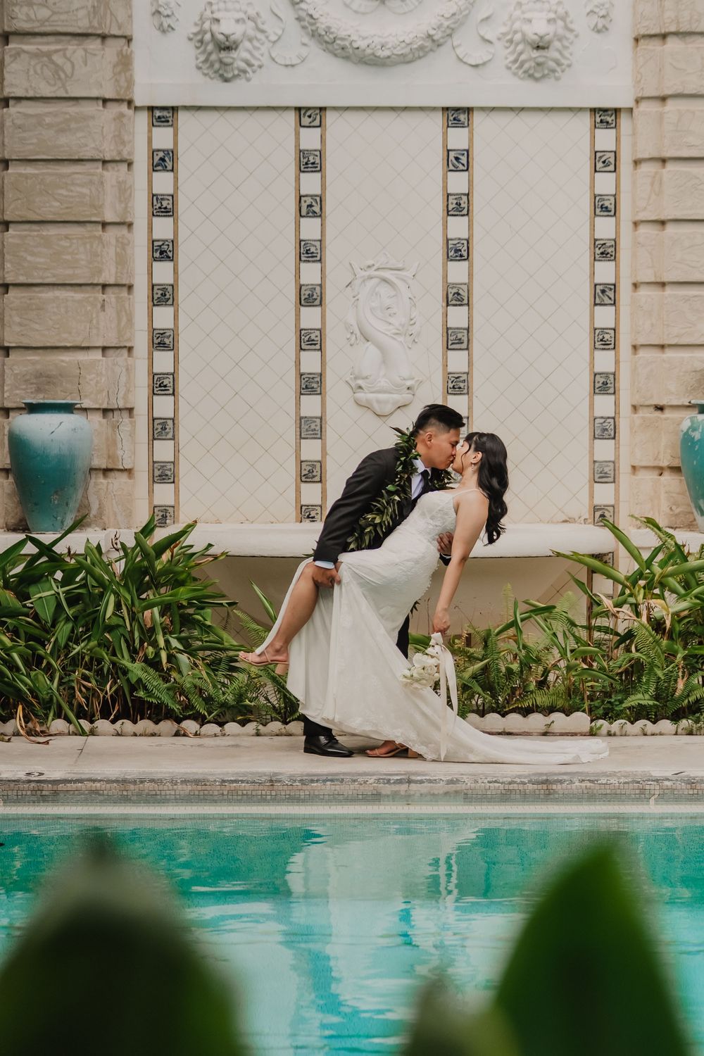 A romantic couple shares a kiss in front of an ornate stone wall with tropical plants and a swimming pool.