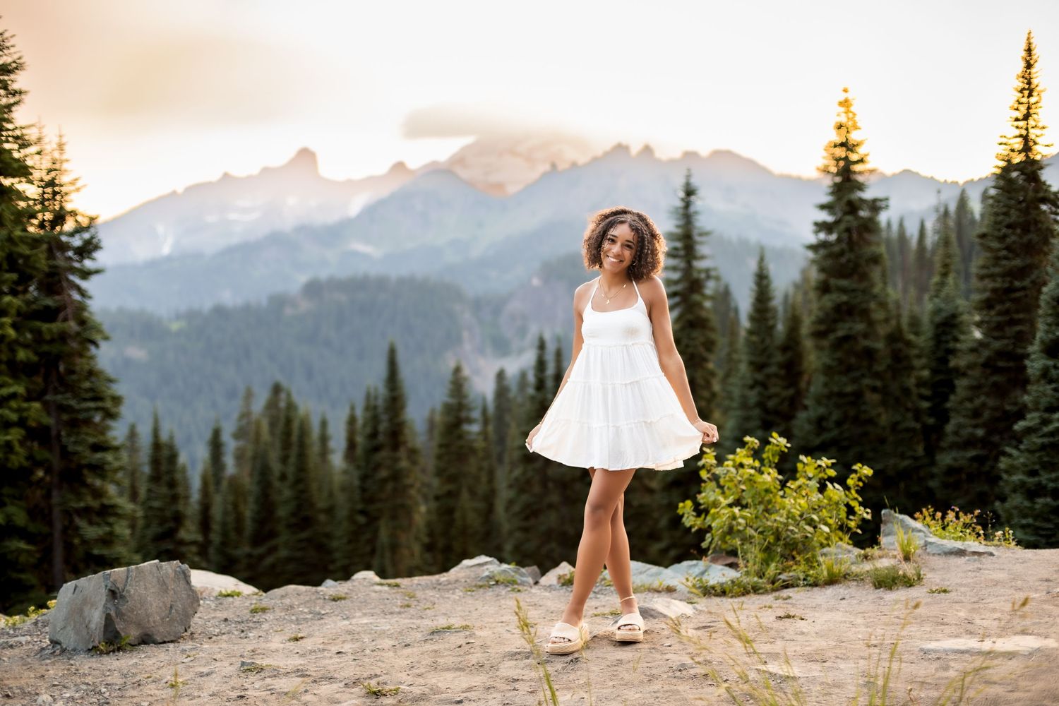 A whimsical pose in a white dress against a backdrop of mountain peaks and evergreen forest.