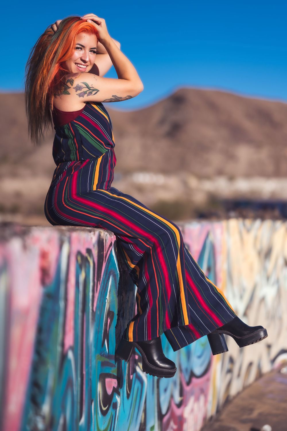 A woman with vibrant orange hair and tattoos poses confidently on a graffiti-covered wall, wearing a striped jumpsuit and black platform boo