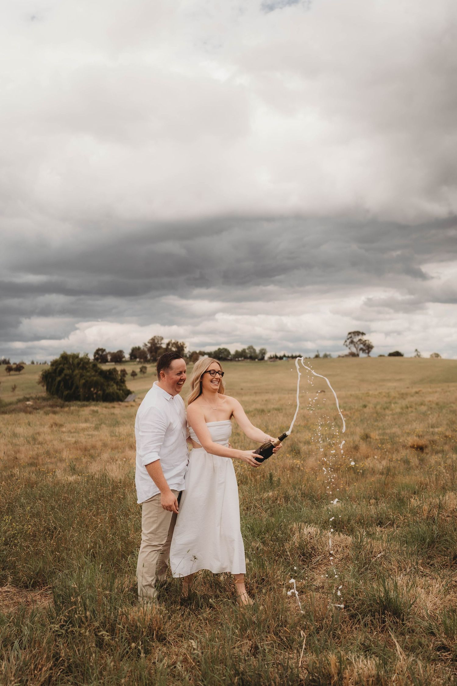 Couple celebrating in a grassy field as champagne sprays from a bottle under cloudy skies.