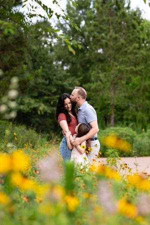 A couple embraces and kisses in a lush garden with yellow wildflowers, while a child hugs them from the front.