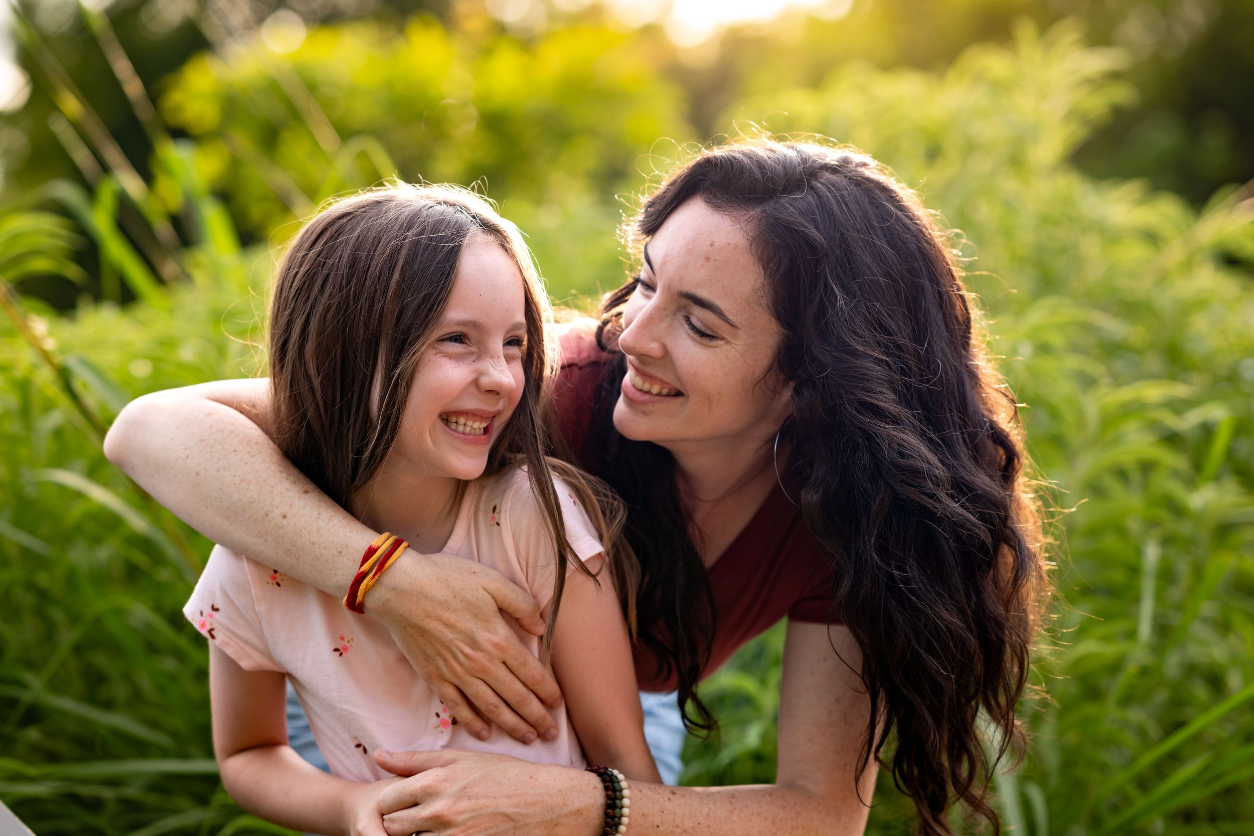 A woman and young girl share a joyful embrace outdoors, smiling at each other among lush green plants.