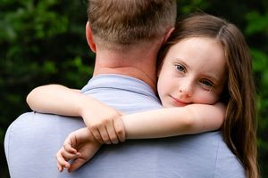Young girl with green eyes hugging a man outdoors, resting her head on his shoulder with green trees behind.