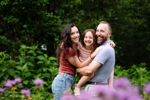 A happy family of three smiling together outdoors surrounded by lush green foliage and purple flowers.