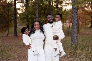 A joyful Black family of four in matching white outfits poses outdoors among autumn trees in a warm portrait.
